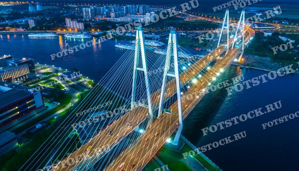 На фотографии изображён вид на Вантовый мост в Санкт-Петербурге в ночное время. Мост освещён многочисленными огнями, создающими живописный световой узор, отражающийся в водах реки. Фотография сделана с высоты, что позволяет оценить великолепие городской инфраструктуры и окружающий городской пейзаж. Это изображение может быть использовано в материалах, посвященных архитектуре, инженерии, городскому планированию, а также в туристических брошюрах и постерах.